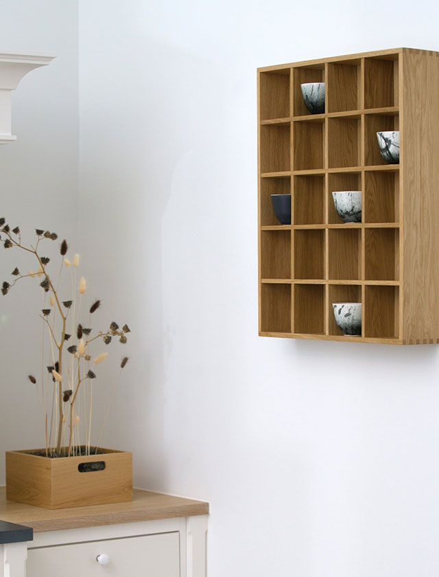 Tassenregal aus Eicheholz von Kath & Andersen, das in einer stilvollen Designküche an der Wand platziert ist. In den quadratischen Fächern des Wandregals sind verschiedene Sammeltassen und Keramikbecher dekorativ einsortiert. Vor dem Regal steht eine Person, die gerade eine Tasse abstellt. Die helle Natursteinwand, goldene Pendelleuchten, ein grüner Küchenschrank und ein frischer Basilikumtopf auf der Arbeitsfläche unterstreichen den hochwertigen und natürlichen Charakter der Szene.“