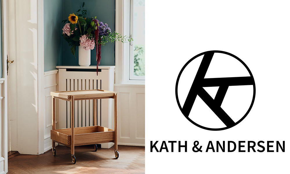 In einem hellen, elegant eingerichteten Raum steht ein minimalistischer Servierwagen aus hellem Holz von Kath & Andersen. Der Wagen ist auf vier goldfarbenen Rollen montiert und besteht aus einem oberen Tablett sowie einer unteren Ablagefläche. Er wirkt leicht, funktional und gleichzeitig edel – typisch für skandinavisches Design. Im Hintergrund steht eine weiße Vase mit bunten Sommerblumen, die dem Raum einen freundlichen, wohnlichen Charakter verleihen. Rechts daneben ist das Logo von Kath & Andersen in Schwarz abgebildet, bestehend aus einem stilisierten Kreis mit den Buchstaben K und A sowie dem Schriftzug „KATH & ANDERSEN“.