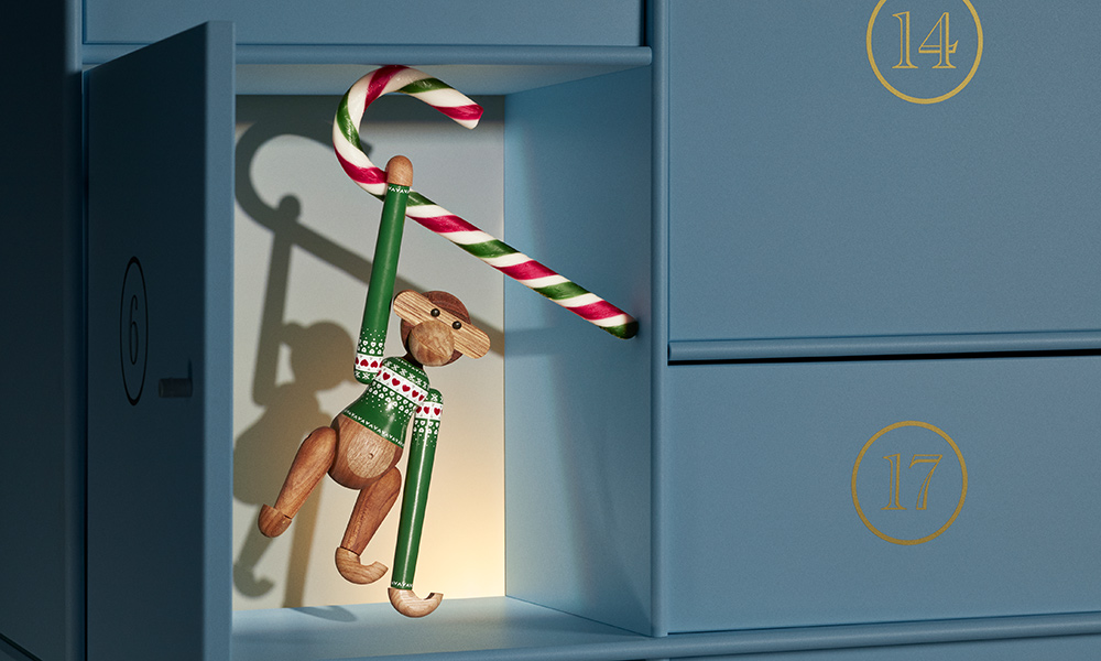 ay Bojesen Affe mit grünem Christmas Jumper hängt an einer rot-weiß-grünen Zuckerstange in einer weihnachtlichen Adventskalender-Szenerie.