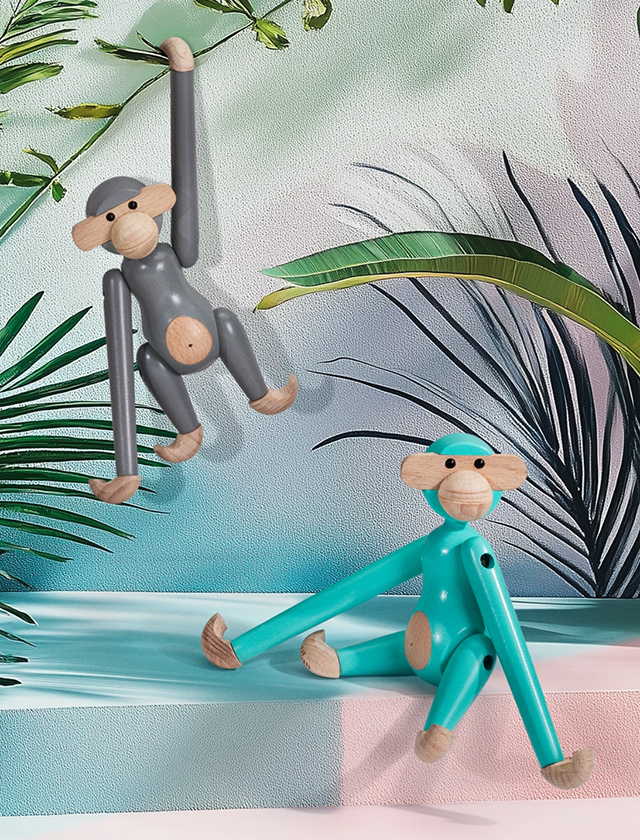 Zwei Kay Bojesen Monkey Affen in Grau und Türkis aus Holz in einer modernen, tropischen Szene: Der graue Affe hängt mit einem Arm nach oben, der türkise sitzt entspannt auf einer Stufe. Im Hintergrund sind dekorative Palmblätter und eine pastellfarbene Wand.