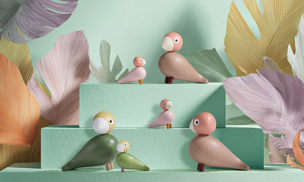 Kay Bojesen Vogelpaar Liv und Lykke in Grün und Rosa auf mintfarbenen Podesten, dekorativ arrangiert vor pastelligem Blattmotiv-Hintergrund.