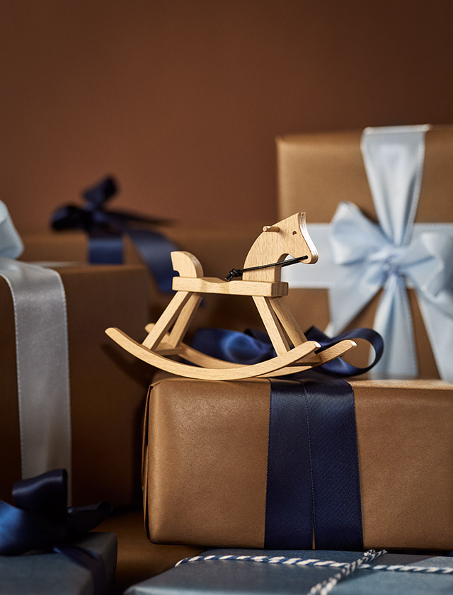 Kay Bojesen Mini-Schaukelpferd aus FSC®-zertifiziertem Buchenholz als dänische Designklassiker auf einem Geschenk stehend vor weiteren Geschänkkisten