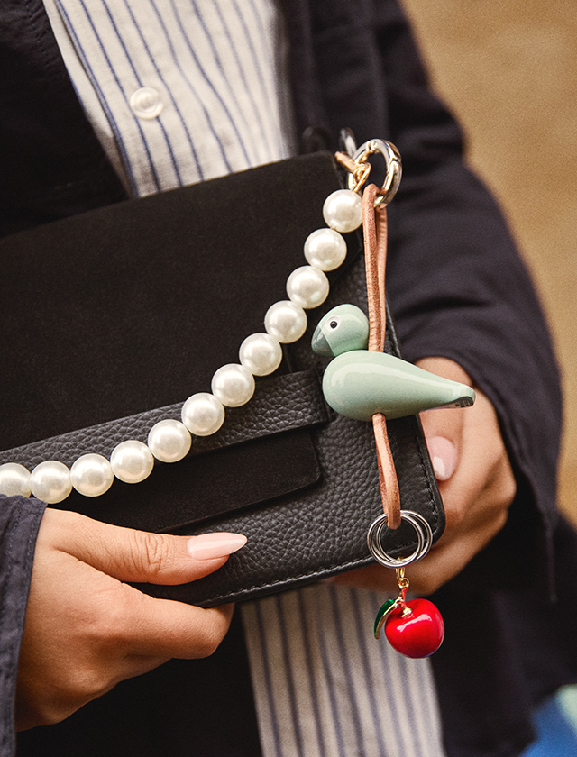 Orangefarbener Kay Bojesen Vogel Schlüsselanhänger hängt an einer grauen Handtasche, getragen in der Hand im Streetstyle-Look Orange an grauer Handtasche – Streetstyle