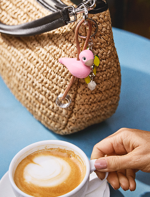 Rosa Kay Bojesen Vogel Schlüsselanhänger an Strohtasche, Detailaufnahme im Café neben einer Tasse Kaffee
