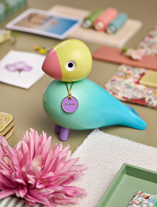 Glänzende Kay Bojesen Story Bird Singvogel „Glæde/Joy“ in Aqua/Türkis mit gelbgrünem Kopf, rosa Schnabel und Token-Anhänger im Lifestyle-Setting.