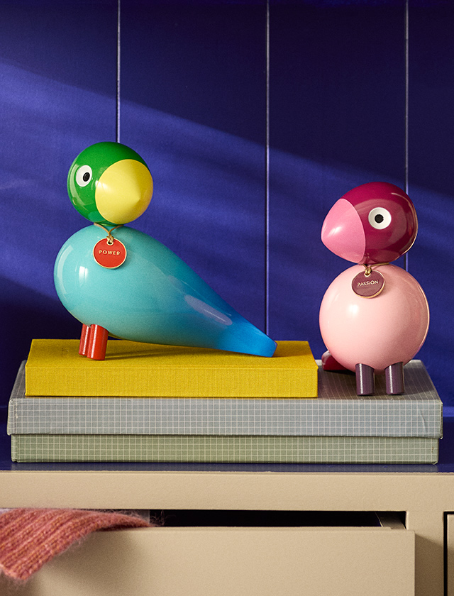 Kay Bojesen Story Bird Power in Blau und Grün dekorativ auf Büchern arrangiert – farbenfrohe Designfigur mit gelbem Schnabel und rotem Anhänger.