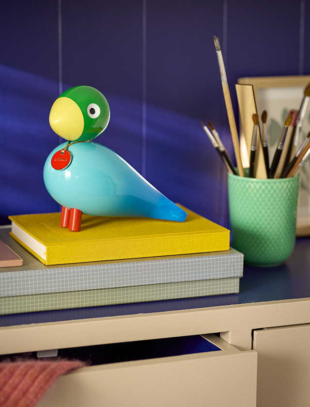Kay Bojesen Story Bird Power in Türkis und Grün auf gelbem Buch neben einem grünen Lyngby Porcelæn Rhombe Color Becher mit Pinseln vor dunkelblauem Hintergrund.