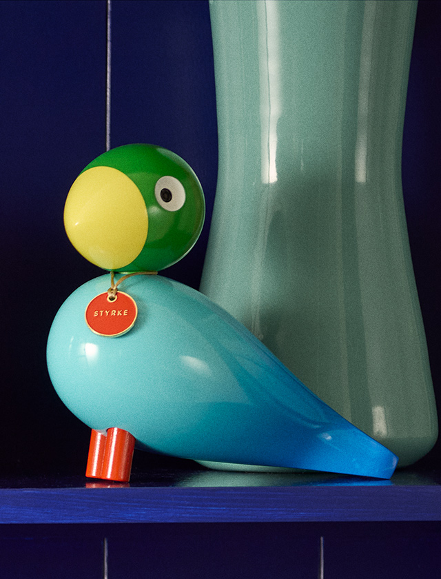 Kay Bojesen Story Bird Power in Türkis, Grün und Gelb neben einer hohen grünen Vase vor dunkelblauem Hintergrund.