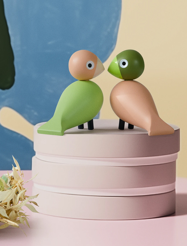 Zwei Kay Bojesen Unzertrennliche aus Holz in Hellgrün und Rosa stehen auf Podesten; eines der Podeste erinnert an eine stilisierte Torte.