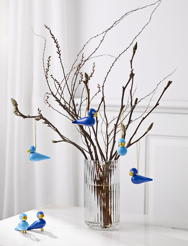 Blaue Kay Bojesen Wintervögel hängen an Zweigen in einer Lyngby Porcelæn Glasvase; ein blaues Wintervogel-Paar steht auf der hellen Marmortischplatte.