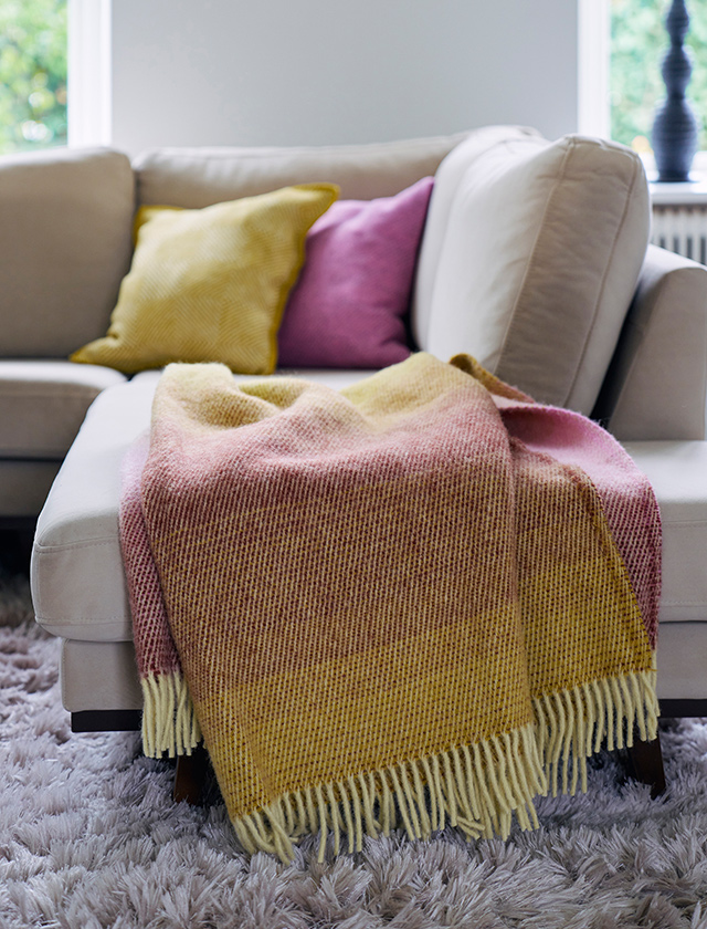 Klippan Yllefabrik Aurora Dawn Wolldecke mit Farbverlauf in Gelb, Rosa und warmen Naturtönen auf einem hellen Sofa mit farblich passenden Kissen.