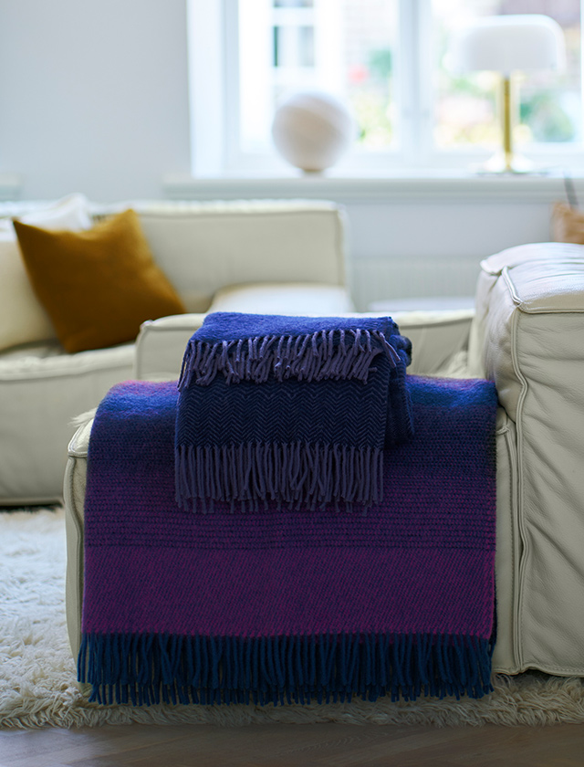 Aurora Wolldecke von Klippan Yllefabrik in kräftigem Magenta mit dunkelblauen Fransen, dekorativ über ein helles Ledersofa gelegt. Die hochwertige Wolldecke setzt stilvolle Farbakzente im Wohnzimmer und kombiniert skandinavisches Design mit zeitloser Eleganz. Perfekt als Sofa- oder Kuscheldecke für gemütliche Stunden.