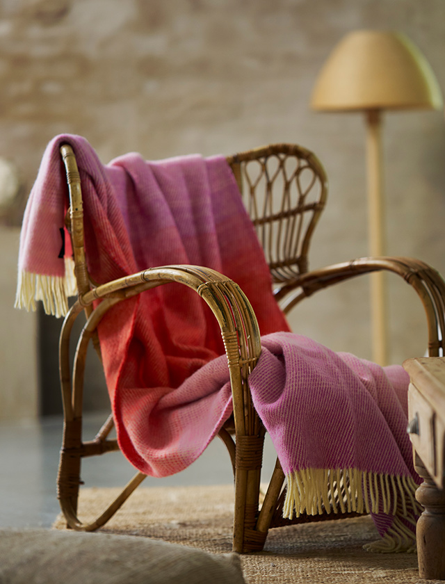 Klippan Yllefabrik Aurora Wolldecke in Reddish Pink mit leuchtendem Farbverlauf in Rosa- und Rottönen, dekorativ über einem Rattansessel im Wohnzimmer.