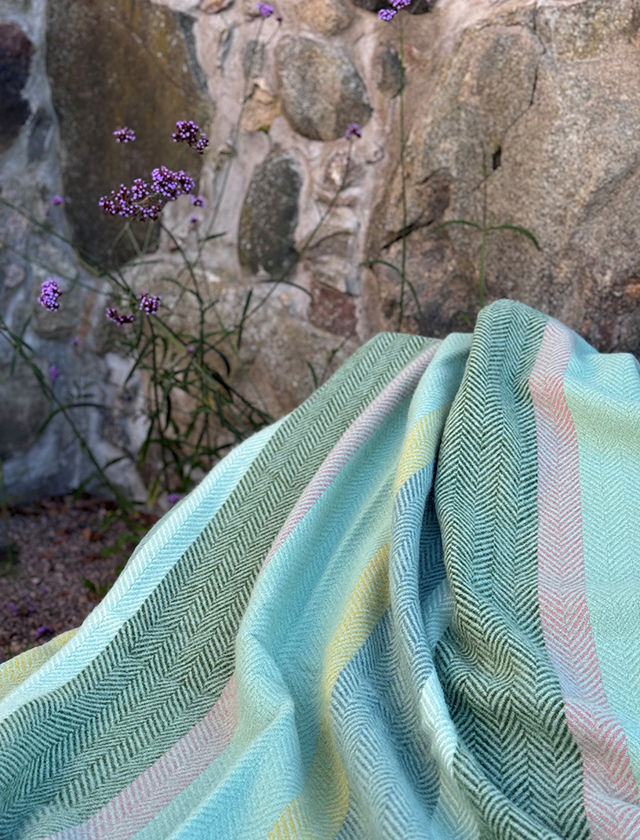 Klippan Yllefabrik Birka Decke in Meadow mit grün-blauen und pastellfarbenen Streifen im Fischgrätmuster, dekorativ im Außenbereich drapiert.
