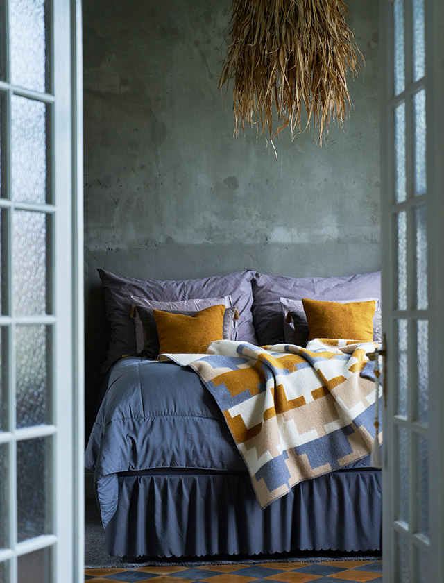 Klippan Cancun Wolldecke mit grafischem Muster in Blau, Senfgelb, Beige und Weiß, dekoriert auf einem Bett mit passenden gelben Gotland Kissen im skandinavischen Schlafzimmer.
