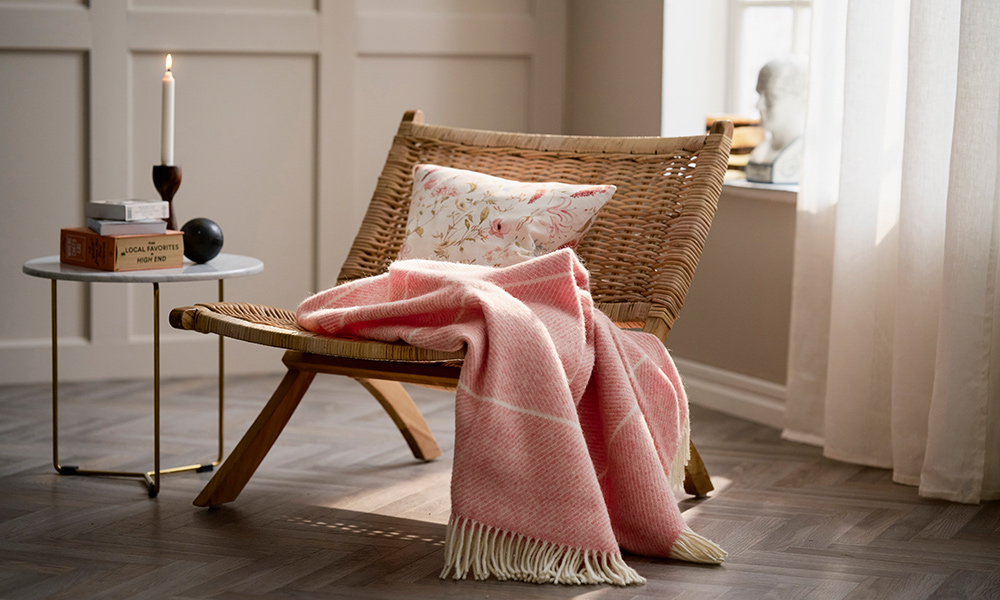 Rosafarbene Klippan Frank Wolldecke in Salmon lässig über einen Korbstuhl drapiert, daneben ein kleiner Beistelltisch mit Kerze in wohnlichem Innenraum.