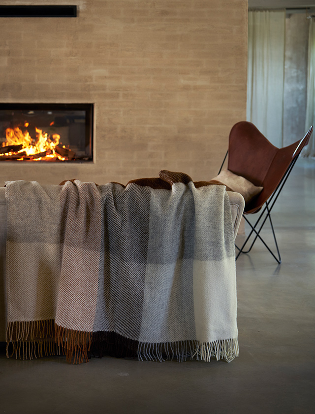 Klippan Yllefabrik Gotland Check Wolldecke in Beige mit großflächigem Karomuster, dekorativ über einem Sofa vor einem Kamin drapiert.