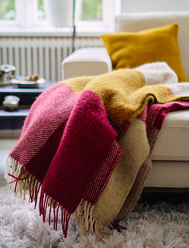Klippan Yllefabrik Gotland Check Wolldecke in Burgundy, Gelb und Naturtönen dekorativ auf einem hellen Sofa mit gelbem Kissen.