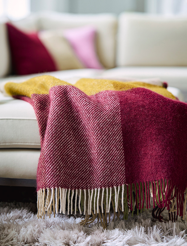 Klippan Yllefabrik Gotland Check Wolldecke in Burgundy, Gelb und Natur mit Fransen, dekorativ über einem hellen Sofa drapiert.