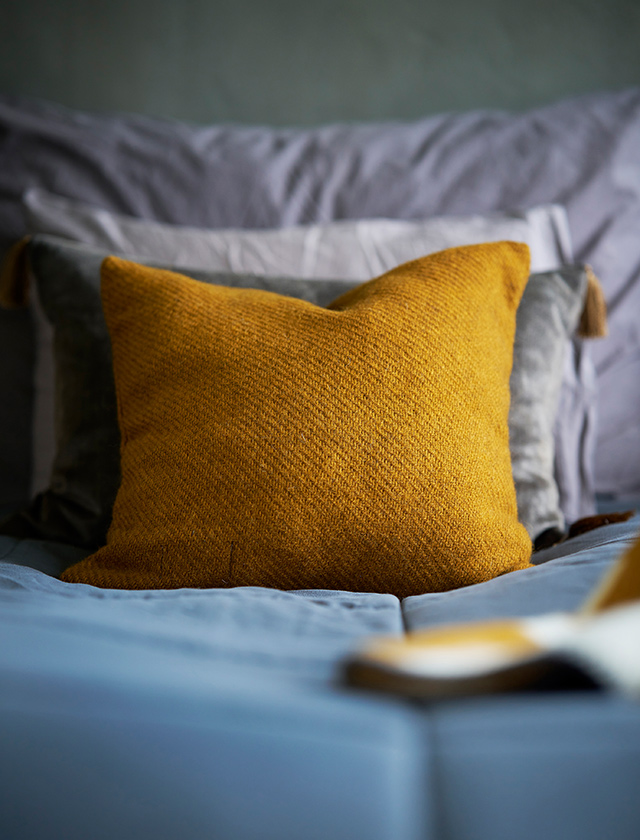 Klippan Gotland Kissenbezug in Caramel mit warmer, strukturierter Wolloberfläche, dekorativ auf einem Bett mit grauen Kissen im skandinavisch eingerichteten Schlafzimmer.