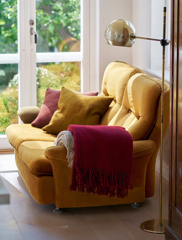 Gemütliche Wohnszene mit einer Klippan Yllefabrik Gotland Wolldecke in Burgundy über einem gelben Sofa, kombiniert mit Kissen in warmen Caramel- und Senftönen. Das Arrangement neben einer großen Fensterfront verbindet skandinavisches Design mit wohnlicher Atmosphäre und zeigt die perfekte Kombination aus hochwertigen Naturmaterialien und harmonischen Farben.