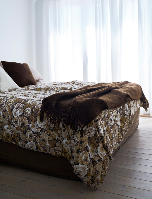 Ein helles Schlafzimmer mit großer Fensterfront, am Bettende liegt eine Klippan Yllefabrik Gotland Wolldecke in der Farbe Walnut. Die Wolldecke aus Gotlandwolle und Lammwolle setzt einen warmen, erdigen Akzent auf der floralen Tagesdecke und sorgt für ein elegantes, skandinavisches Ambiente.