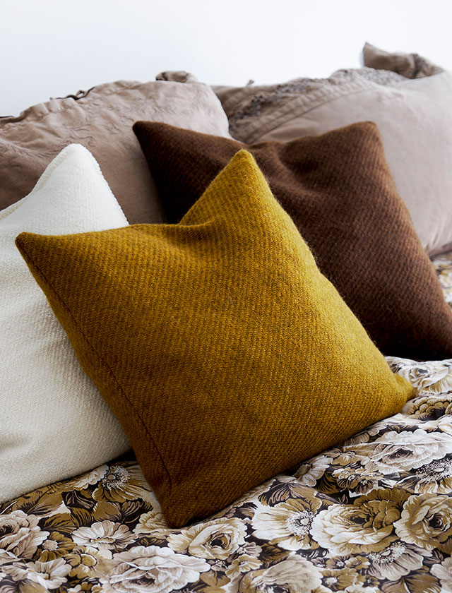 Wollkissenbezüge der Serie „Gotland“ von Klippan Yllefabrik. Die Kissenbezüge in Cremeweiß, Senfgelb und Dunkelbraun sind aus hochwertiger Gotland-Wolle gefertigt und liegen auf einem floral gemusterten Bettbezug in warmen Erdtönen. Die natürliche Textur der Wolle und die warmen Farben unterstreichen die skandinavische Wohnästhetik und schaffen eine gemütliche, einladende Atmosphäre. Klippan Yllefabrik steht für nachhaltige Heimtextilien aus reiner Schurwolle, gefertigt in traditioneller Webkunst mit Fokus auf Naturmaterialien und Langlebigkeit.