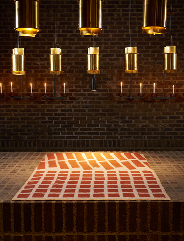 St Petri Kyrka Floor Wolldecke von Klippan Yllefabrik, hier in einer markanten Variante mit rotem, architektonisch inspiriertem Muster auf hellem Grund. Die Decke liegt vor einer stimmungsvollen Backsteinwand mit goldenen Leuchten und Kerzen, was die klare, grafische Gestaltung und den besonderen Charakter dieses hochwertigen Wohntextils unterstreicht.