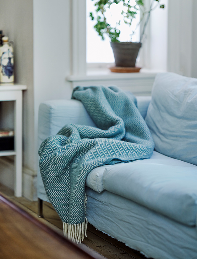 Eine hellblaue Sorbet Wolldecke von Klippan Yllefabrik liegt dekorativ über einem Sofa im Wohnzimmer. Die Decke aus 100 % Bio-Wolle zeigt ein feines Zickzackmuster mit hellen Fransen und bringt skandinavische Leichtigkeit und Behaglichkeit in den Raum. Sie verbindet nachhaltige Materialien mit zeitlosem Design – perfekt als Sofadecke, Wohnaccessoire oder zum Einkuscheln.