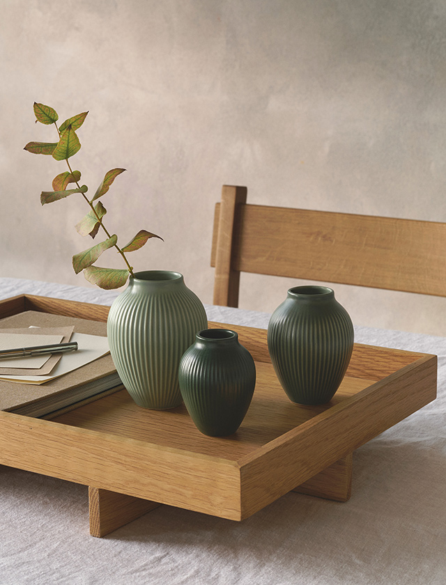 Drei Miniatur-Rillenvasen von Knabstrup Keramik in den matten Grüntönen Misty Green, Celadongrün und Dark Green stehen dekorativ auf einem Tablett aus Holz. Das markante Rillendekor betont die elegante Form der kleinen Vasen und unterstreicht ihre skandinavische Schlichtheit. Ob mit einzelnen Zweigen bestückt oder pur arrangiert – die Miniaturvasen setzen stilvolle Akzente und eignen sich perfekt als feines Designobjekt für Tisch, Sideboard oder Regal.