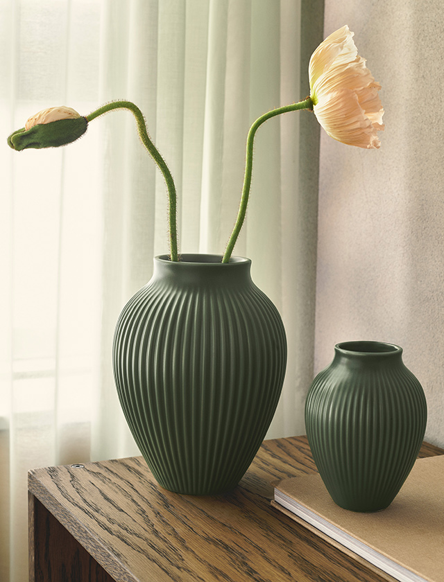 Zwei Knabstrup Keramik Rillenvasen in matter Celadongrün-Glasur stehen dekorativ auf einem Holztisch vor einem lichtdurchfluteten Vorhang. Die Kombination aus großer und kleiner Vase betont die elegante Form mit vertikaler Rillenstruktur und verleiht den Vasen skandinavische Klarheit. Mit oder ohne Blumenarrangement lassen sich die Vasen als stilvolle Designobjekte einsetzen und setzen natürliche, zeitlose Akzente im Wohnraum.