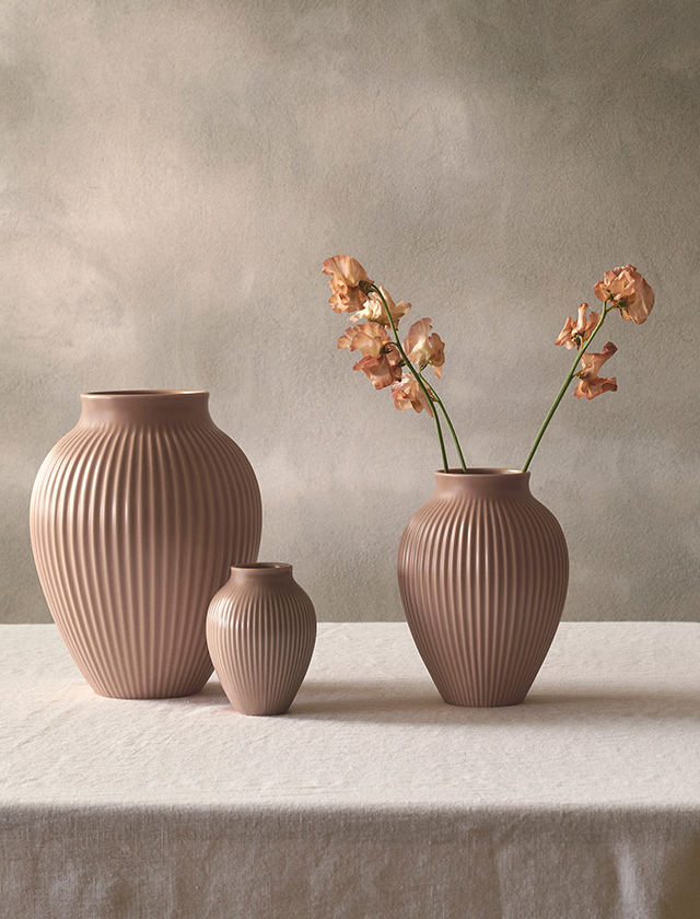 Drei Knabstrup Keramik Rillenvasen in der eleganten Farbvariante Dusty Rose stehen dekorativ auf einem Tisch mit Leinentuch. Das matte Rosé mit pudrigem Unterton erinnert an verblühte Rosenblätter und Abendlicht und verleiht den Vasen eine ruhige, zeitlose Ausstrahlung. Die charakteristische vertikale Rillenstruktur betont die organische Form und sorgt für elegante Dynamik. In drei Größen arrangiert, lassen sich die Vasen vielseitig einsetzen – als edler Rahmen für Blumenarrangements oder als skulpturale Designobjekte im skandinavischen Wohnstil.