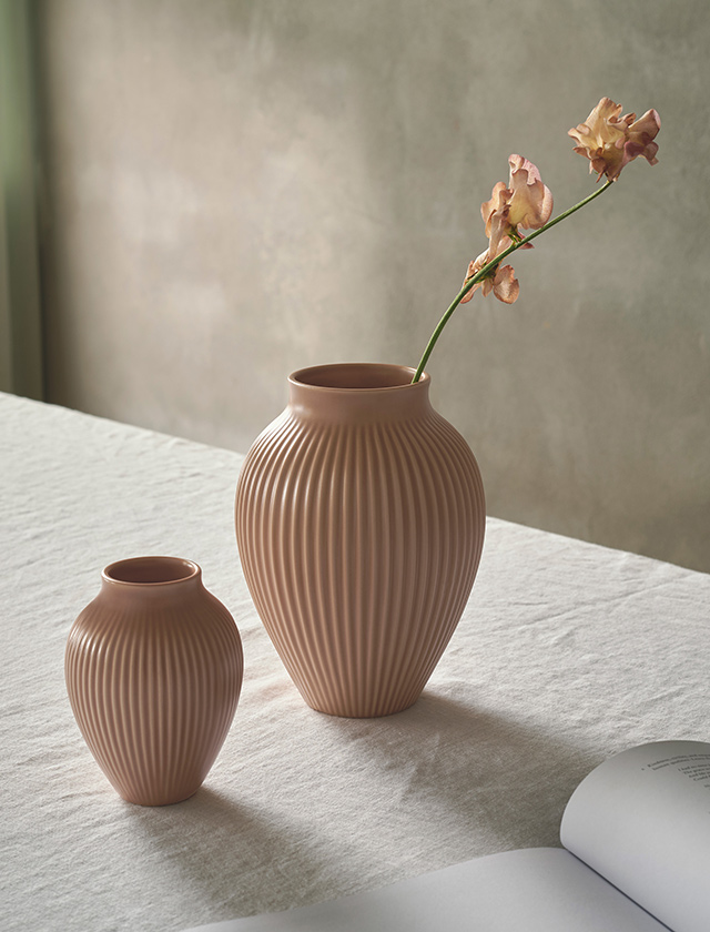 Zwei Knabstrup Keramik Rillenvasen in matter Dusty Rose Glasur stehen dekorativ auf einem hellen Tisch. Die elegante vertikale Rillenstruktur betont die organische Form der Vasen und verleiht ihnen skandinavische Schlichtheit. Arrangiert mit einer zarten Blüte oder als puristische Designobjekte setzen die Vasen stilvolle Akzente und fügen sich harmonisch in moderne wie klassische Interieurs ein.