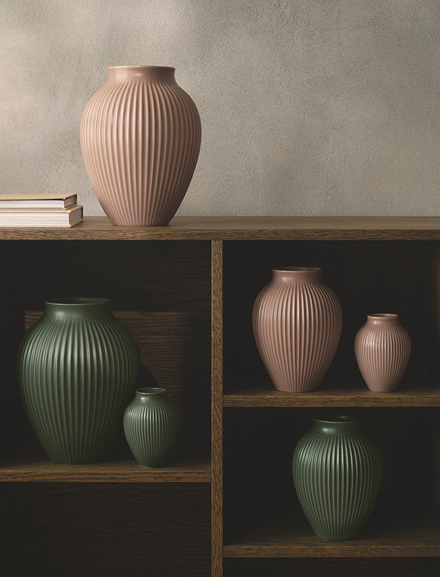 Knabstrup Keramik Rillenvasen in den matten Farbtönen Dusty Rose und Grün (Celadongrün und Dark Green) sind dekorativ in einem offenen Holzregal arrangiert. Die markante vertikale Rillenstruktur betont die elegante Form der Vasen und verleiht ihnen zeitlose Ausstrahlung. Ob einzeln aufgestellt oder in Kombination, die Vasen der Serie setzen stilvolle Akzente im skandinavischen Wohnstil und eignen sich sowohl für Blumenarrangements als auch als reine Designobjekte.
