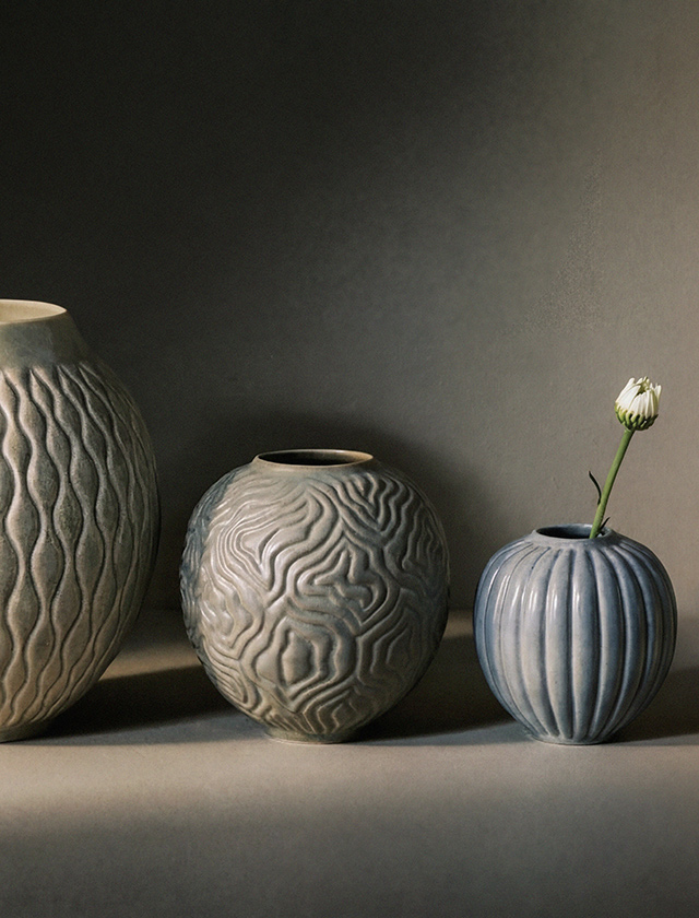 Die größere Vase enthält mehrere violett blühende Zierlauch-Blumen (Allium), deren kugelförmige Blüten einen schönen Kontrast zur dunklen Vase und der hellen Umgebung bilden. Die mittlere Vase steht daneben und bleibt leer, wodurch die Form und das Design der Vasen noch besser zur Geltung kommen.