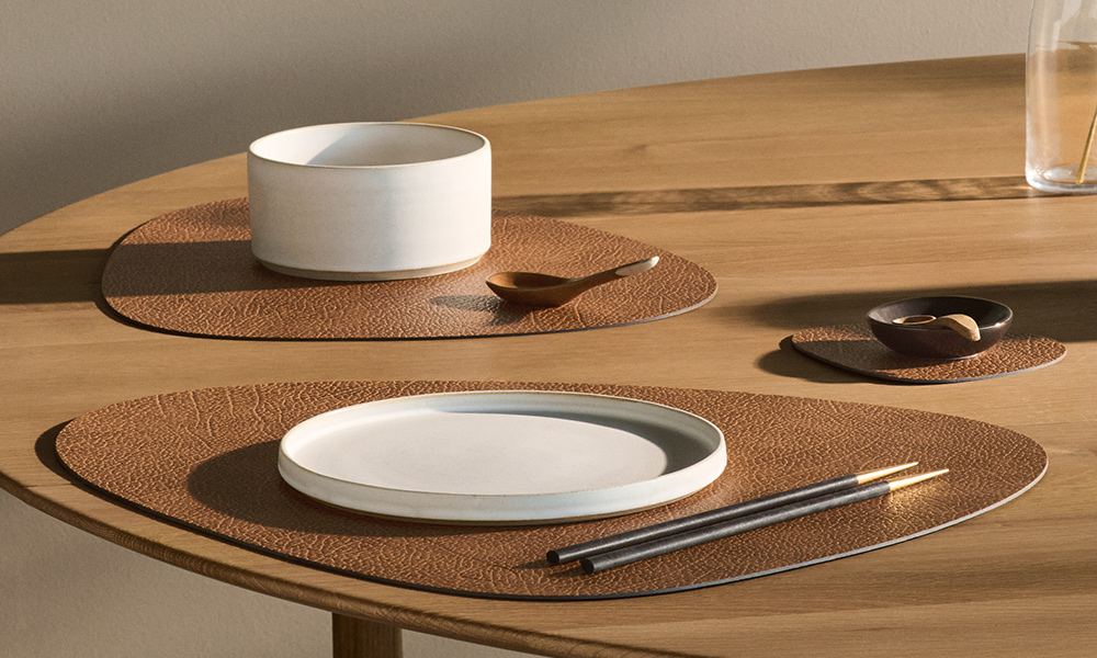 Tischarrangement mit den charakteristischen Tischsets von LIND DNA in einem warmen Braunton. Die organisch geformten Platzsets aus hochwertigem, recyceltem Leder fügen sich harmonisch in die natürliche Ästhetik des skandinavischen Designs ein. Auf dem hellen Holztisch sorgen die weichen Formen und die fein strukturierte Lederoberfläche für ein besonders edles, ruhiges Gesamtbild.