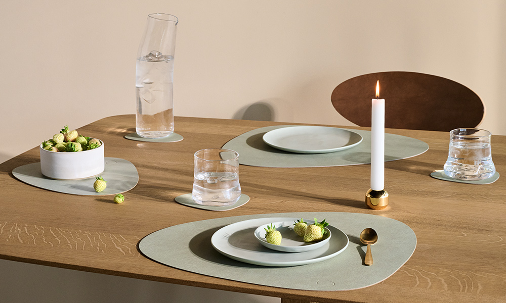 Elegante LIND DNA Curve Tischsets aus recyceltem Leder in einem modernen, minimalistischen Ambiente. Die hochwertigen Platzsets in sanften Naturtönen unterstreichen das skandinavische Designkonzept und setzen zusammen mit dem klaren Geschirr, der schlichten Kerze und den weißen Erdbeeren ein zeitloses Statement für nachhaltige Tischkultur. Die Kombination aus Ästhetik und Umweltbewusstsein macht die LIND DNA Curve Tischsets zur idealen Wahl für designaffine Haushalte, die auf Qualität und Nachhaltigkeit setzen.