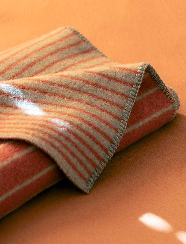 Die AURORA-Decke von Lapuan Kankurit liegt locker gefaltet auf einer warm getönten, zimtorangen Oberfläche. Das Design zeigt ein feines Streifenmuster in natürlichem Zimtorange und hellem Beigegrau. Der Rand ist mit einem dekorativen Überwendlingsstich eingefasst. Die Decke wurde mit natürlichen Farbstoffen gefärbt, darunter Waid (woad). Einfallendes Sonnenlicht bringt die weiche Wollstruktur und die sanften Farbtöne stimmungsvoll zur Geltung.