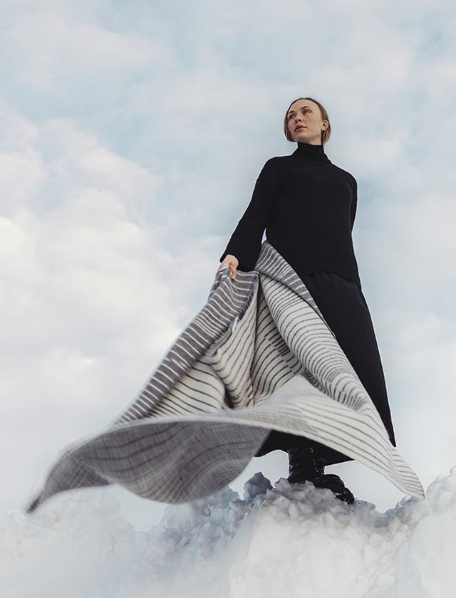 Die Aurora Wolldecke von Lapuan Kankurit in Grau-Weiß weht dramatisch im Wind, während eine Frau auf einem schneebedeckten Hügel steht – stilvoll, minimalistisch und nordisch inspiriert. Ideal für hochwertige Winter-Content-Kampagnen.