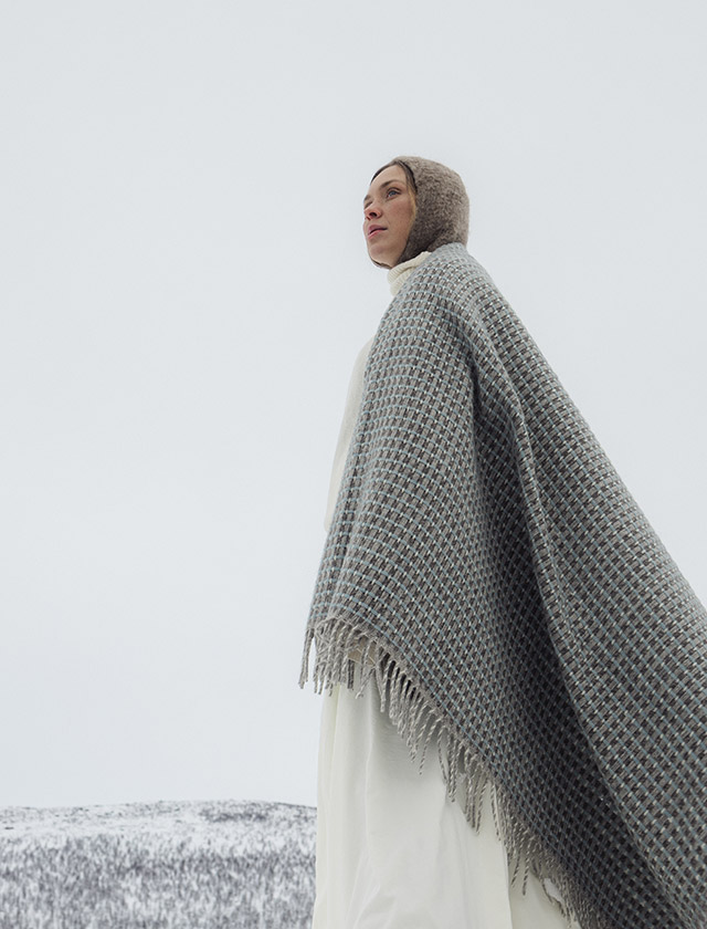 Havas Wolldecken von Lapuan Kankurit aus 100 % Wolle, gefertigt aus weicher finnischer Finnschafwolle mit charakteristischem geometrischem Webmuster. Die Decke in der Farbe Natural-Dyed-Woad ist wie ein Umhang über die Schultern einer Frau gelegt und unterstreicht ihre skandinavische Ausstrahlung.