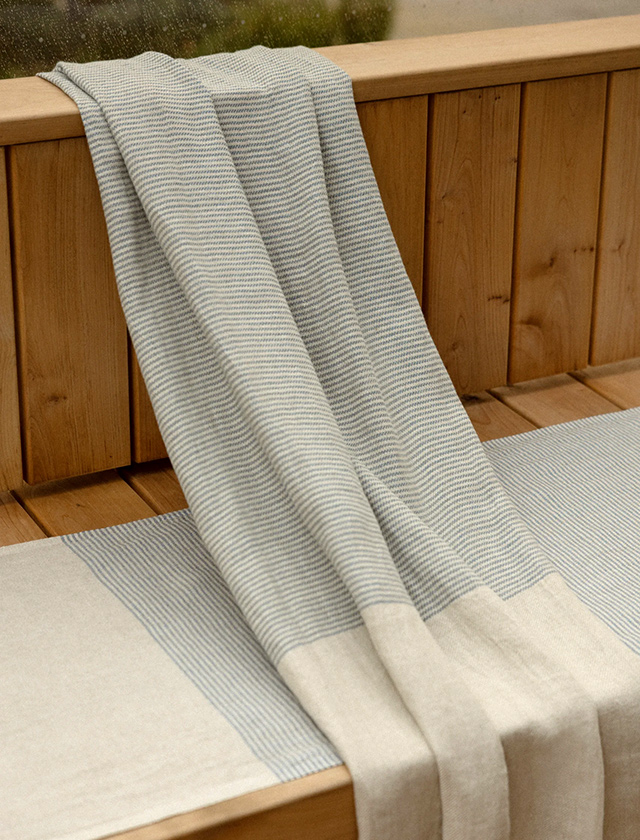 Ein Mann steht entspannt in einer traditionell eingerichteten Sauna, umhüllt von einem stilvollen Frottiertuch im Birken-Design „Koivu“ der finnischen Marke Lapuan Kankurit. Das hochwertige Saunatuch aus Leinen-Baumwoll-Mischgewebe ist ideal für Wellness- und Saunagänge. Die dezente Farbgebung in Grautönen kombiniert mit dem skandinavisch inspirierten Birkenmuster sorgt für eine natürliche und zugleich elegante Atmosphäre. Im Hintergrund sieht man einen Saunaofen, Dampf und einen klassischen Saunaeimer – perfekte Details für authentische Wohlfühlmomente.