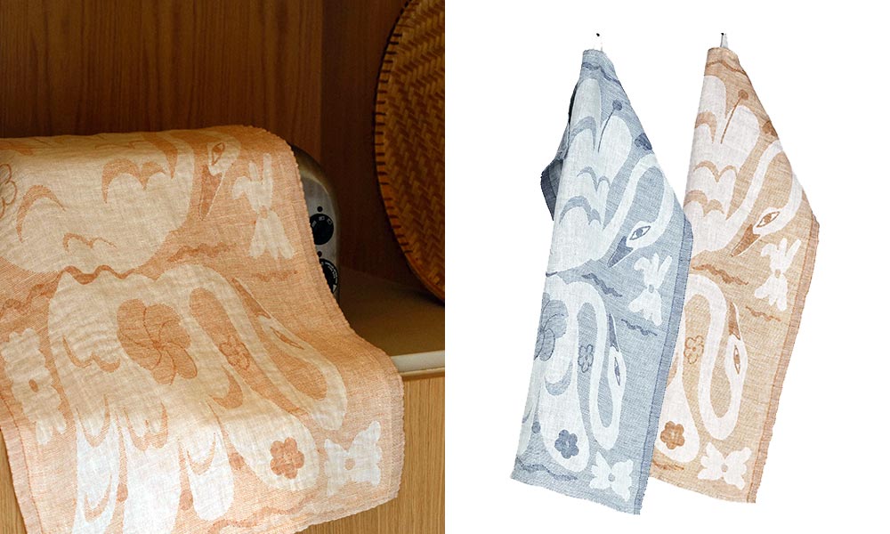 Zwei stilvolle Leinentücher der Serie „Kaksin“ von Lapuan Kankurit, aufgehängt nebeneinander in den Farben White-Cinnamon und White-Blue. Das Design zeigt verspielte florale Motive und anmutige Schwäne. Links ist das Kaksin Geschirrtuch in Zimt-Weiß dekorativ auf einem Holzregal drapiert, rechts hängen beide Farbvarianten nebeneinander. Hochwertiges skandinavisches Design trifft auf nachhaltige Naturmaterialien.