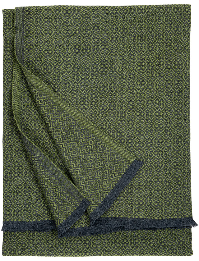 Koli Wolldecke von Lapuan Kankurit, ebenfalls aus 100% Merinowolle. Diese Decke ist in den Farben Schwarz und Braun gehalten und hängt über einem umgestürzten Baumstamm in einem Wald. Die warme Farbkombination und das dichte Webmuster passen perfekt zur natürlichen Umgebung. Die Decke scheint weich und einladend zu sein, ideal für gemütliche Momente in der Natur. Die Sonnenstrahlen, die durch die Bäume fallen, beleuchten die Decke und betonen die natürliche Schönheit und Qualität des Materials.