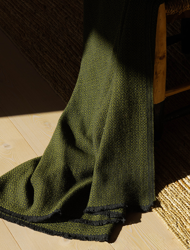Lapuan Kankurit KOLI Merino-Wolldecke in Schwarz und Olive mit grafischem Muster, dekorativ auf hellem Holzboden drapiert.