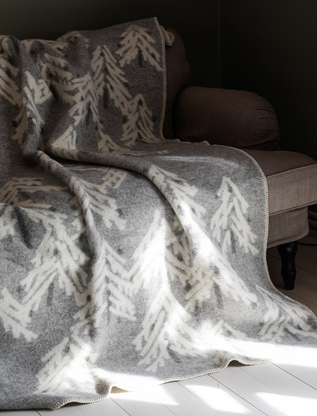Lapuan Kankurit KUUSI Wolldecke in Light Grey-White mit weißem Tannenbaum-Muster, dekorativ über einem Sofa drapiert.