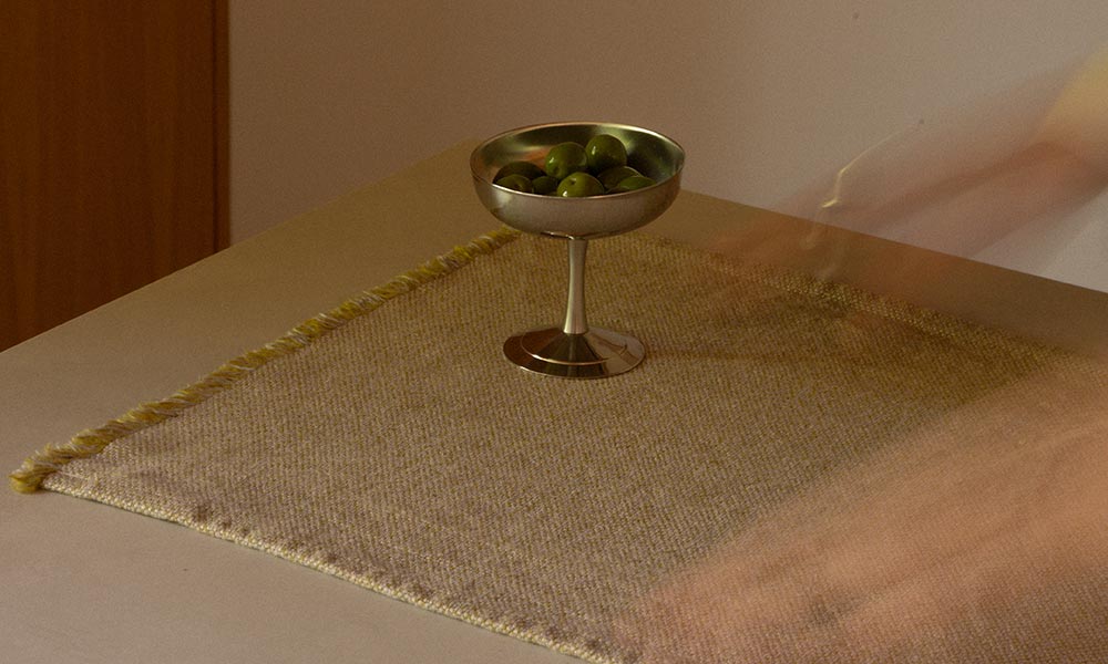 Lounas Tischset von Lapuan Kankurit in der Farbe Olive zu sehen – gewebt aus 100 % Leinen im traditionellen Jacquard-Verfahren. Das Tischset liegt auf einem hellen Tisch und bildet die stilvolle Unterlage für eine schlichte, silberfarbene Schale mit frischen grünen Oliven. Die dezente Webstruktur, die naturbelassene Farbgebung und die feinen Fransen am Rand spiegeln die handwerkliche Qualität und die zeitlose Ästhetik des Produkts wider. Der Schatten einer sich bewegenden Hand im Bild vermittelt ein Gefühl von Alltag, Bewegung und bewusster Tischkultur.