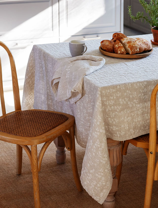 Die Lapuan Kankurit Tischdecke im Design Meirami besteht aus hochwertigem, naturfarbenem 100 % Leinen. Links im Bild liegt sie elegant auf einem Tisch in einem hellen, freundlich gestalteten Essbereich, ergänzt durch frisches Gebäck, eine Tasse und ein passendes Leinentuch. Das feine, eingewebte Kräutermuster in Weiß verleiht dem Gewebe eine zeitlose, skandinavische Note. Rechts ist eine Detailaufnahme zu sehen, die die sorgfältige Umrandung, die Wendbarkeit sowie die klar erkennbare Jacquard-Webstruktur zeigt.
