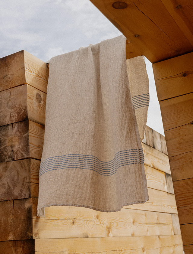Das Lapuan Kankurit Duschtuch „Puro“ aus hochwertigem Leinen–Tencel–Baumwolle in der Farbe leinen-blau hängt locker über einer massiven Holzsauna im Außenbereich. Die natürliche Leinenstruktur und die feinen blauen Streifen verleihen dem Tuch eine zeitlose, skandinavische Ästhetik. Das warme Holz und der helle Himmel unterstreichen den nordischen Charakter – ideal für Sauna, Spa, Wellness und nachhaltige Badkultur. Ein hochwertiges finnisches Designprodukt für stilvolle Bade- und Saunamomente.