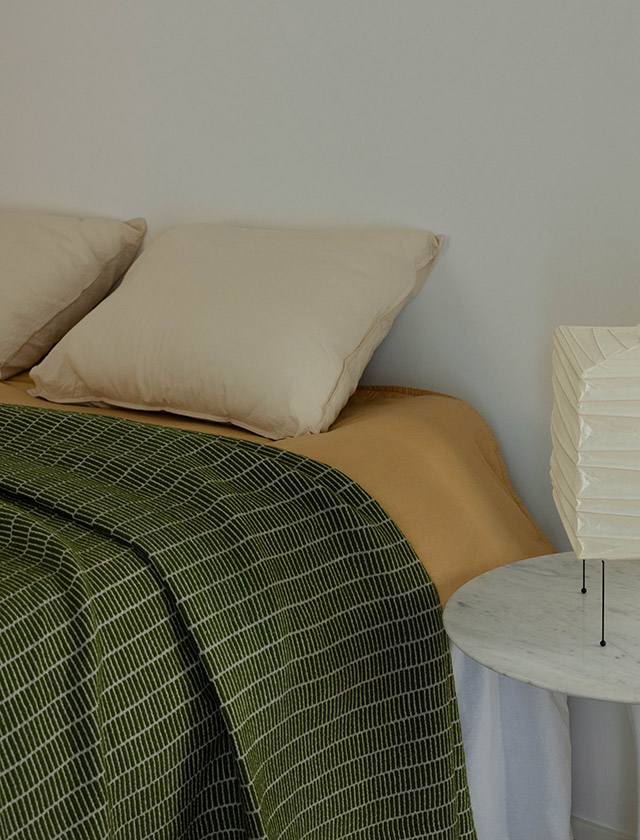 Raitti Tagesdecke von Maija Fagerlund in der Farbe Olive. Die grafisch-linearen Muster orientieren sich an traditionellen finnischen Webtechniken wie der Ripsbindung, inspiriert von Raanu-Wandbehängen und Ripsi-Teppichen. Die Decken bestehen aus 100 % lokaler Finnsheep-Wolle, die für ihre natürliche Weichheit und Wärme bekannt ist.