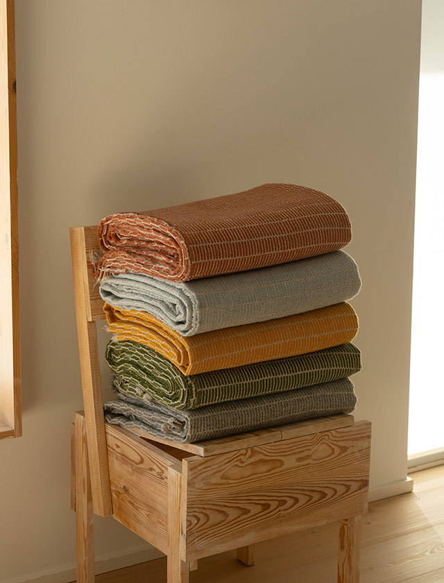 Raitti Tagesdecken von Maija Fagerlund zusammengelgt und als Stapel in den Farben Grey,  Olive, Coffee Dyed Orange, Light-Blue und Cinnamon. Die grafisch-linearen Muster orientieren sich an traditionellen finnischen Webtechniken wie der Ripsbindung, inspiriert von Raanu-Wandbehängen und Ripsi-Teppichen. Die Decken bestehen aus 100 % lokaler Finnsheep-Wolle, die für ihre natürliche Weichheit und Wärme bekannt ist.